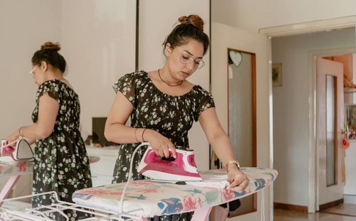 Youngwoman wearing glasses and summer silk dress with steam iron, ironing board in bed room at home in sunny day
