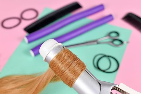 Strand of blond hair wrapped around curling iron with hair styling tools in blurry background,