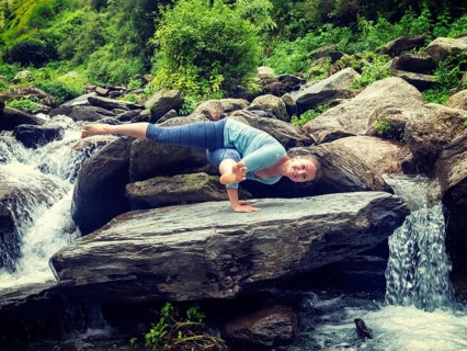 Vintage retro effect hipster style image of sporty fit woman doing yoga asana Eka Pada Koundinyasana 1 at tropical waterfall