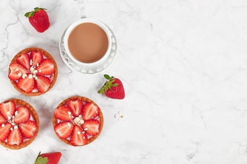 Small tartlet pastries topped with strawberry fruits and white chocolate sprinkles next to coffee cup and copy space