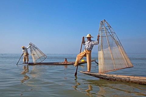Two Intha fishermen steer traditional fishing boats by wrapping their leg around the oar