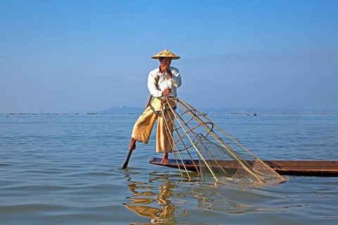 Intha fisherman steering traditional fishing boat by wrapping his leg around the oar
