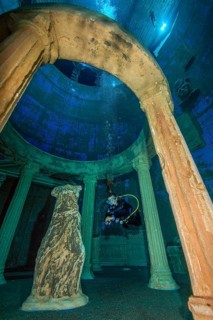 Diver dives through stylised ruin of antique temple round temple in 20 metres water depth in indoor diving tower