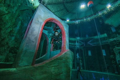 Female diver dives in 10 metres depth in indoor diving tower through imitated bridge house over bow of stylised small ship