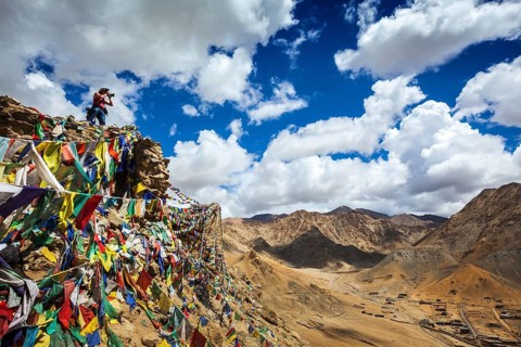 Travel photographer taking photos in Himalayas mountains on cliff with Buddhist prayer flags. Leh