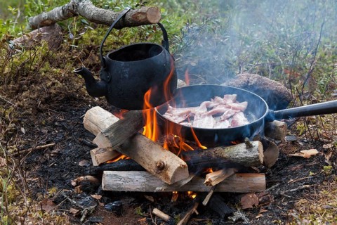 Blackened tin kettle boiling water and pan cooking bacon over flames from campfire