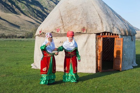 Two Kazakh woman in traditional dress in front of a yurt greeting guests with sweets