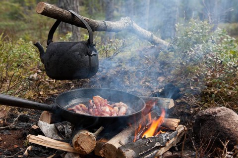 Blackened tin kettle boiling water and pan cooking bacon over flames from campfire