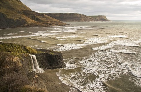 View of coastline with waterfall flowing over hard shale to beach on coast with rough sea