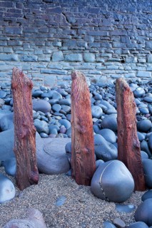 Remains of ancient sea defences emerge from pebbles and shingle in front of modern sea wall on beach