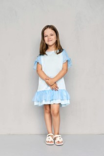 Cute little girl in blue dress and white sandals posing with hands clasped down near the gray wall
