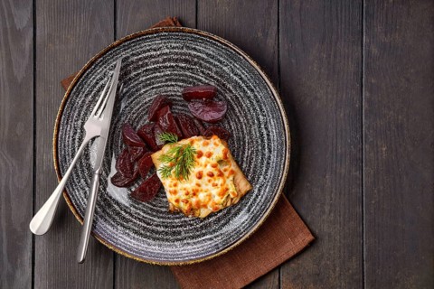Top view of fried cod fillet with melted cheese topping and roasted beetroot slices on dark wooden table