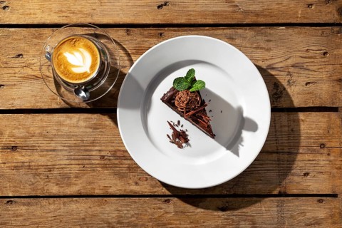 Overhead view of piece of chocolate cake with cappuccino on wooden table under harsh sunlight