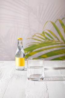 Empty glass on the table and small bottle of tonic water in background under morning sunlight