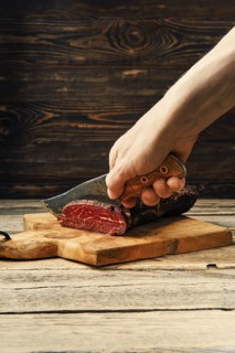 Hand with knife cutting smoked dried beef meat on slices on wooden cutting board