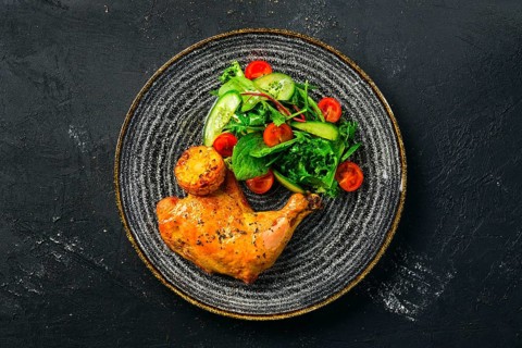 Top view of baked chicken thigh served with fresh vegetables and grilled corn on dark background
