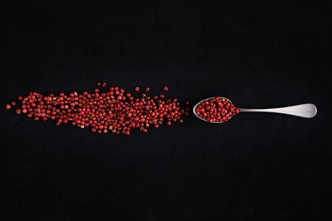 Dry pink pepper scattered near metal spoon on dark background