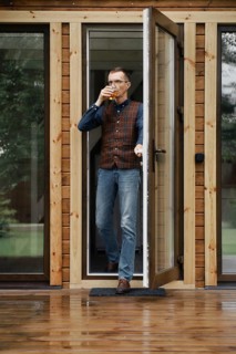 Stylish middle age man comes out on the terrace of a small wooden house with a glass in hand