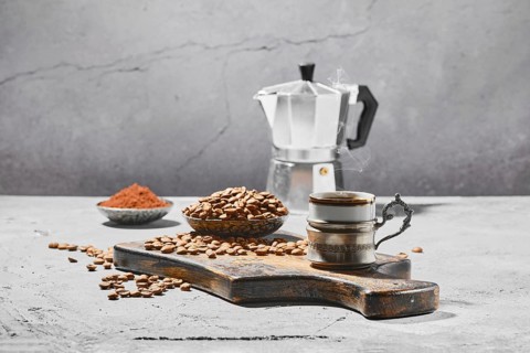 Hot cup of espresso with coffee beans scattered on wooden board and a pot under hard morning sun
