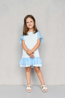 Cute little girl in blue dress and white sandals posing with hands clasped down near the gray wall