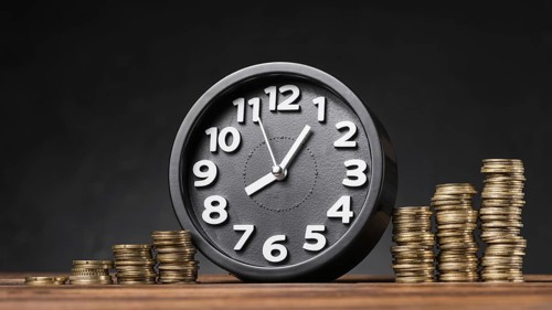 Round clock between the increasing coins on wooden desk against black backdrop
