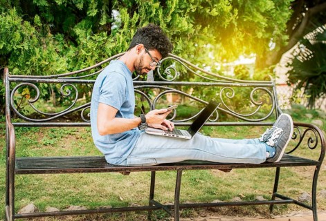 Freelancer man sitting in a park using laptop and cellphone. Man in a park working online with laptop. Relaxed man working with laptop outdoor