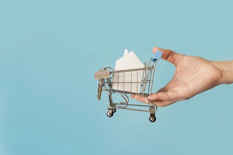 Close up hand holding miniature shopping cart with paper house keys against blue background
