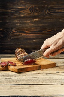 Hand with knife cutting smoked dried beef meat on slices on wooden cutting board