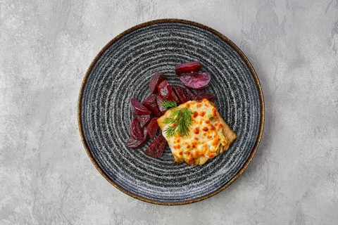 Top view of fried cod fillet with melted cheese topping and roasted beetroot slices on grsy stone background