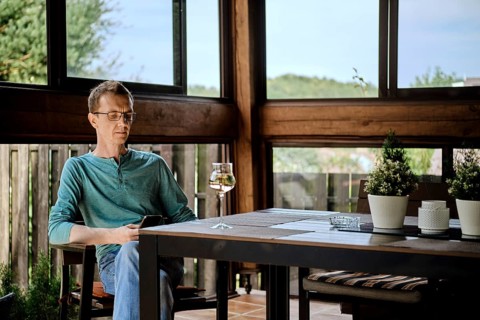 Middle aged man sits on the veranda of country house with a glass of wine and uses smartphone