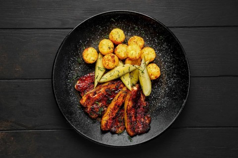 Top view of roasted pork belly with potato balls and pickled cucumber on a plate