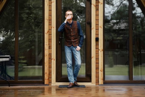 Pensive middle age man stands on the terrace of a wooden bungalow and drinks bourbon