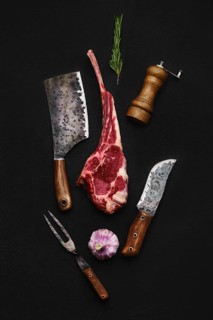 Overhead composition with raw beef cowboy steak and butcher tools over back background
