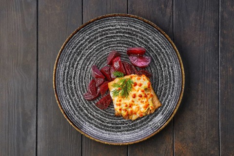 Top view of fried cod fillet with melted cheese topping and roasted beetroot slices on dark wooden table