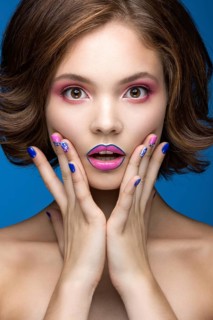 Beautiful model girl with bright makeup and colored nail polish. Beauty face. Short colorful nails. Picture taken in the studio on a blue background