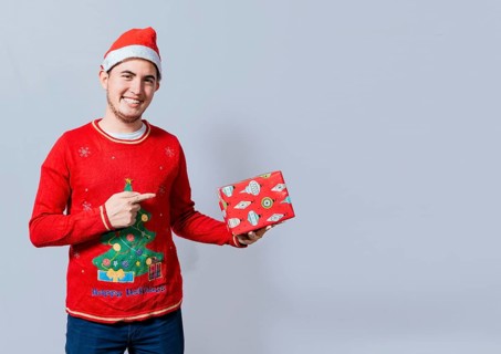 Smiling guy in christmas hat holding and pointing a gift box. Portrait of teenager in christmas hat holding and pointing a gift box. Guy in christmas hat and sweater holding a gift box isolated