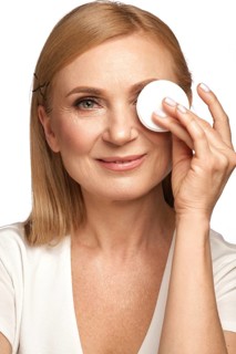 Portrait of a beautiful elderly woman in a white shirt removing makeup with a cleanser and a cotton pad