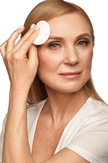 Portrait of a beautiful elderly woman in a white shirt removing makeup with a cleanser and a cotton pad