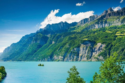 Sunny view over Lake Walen with the small chive island in the turquoise water at the foot of the Churfirsten and the mountain massifs Schaeren and Leistchamm in the background