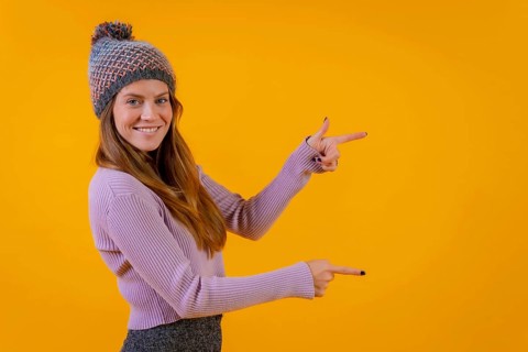 Woman with wool cap on a yellow background pointing to the right smiling