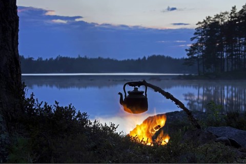 Kettle for making coffee heating over open campfire along lake at sunset