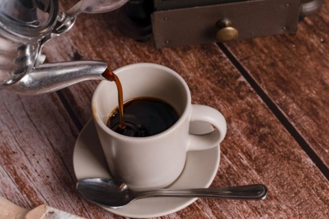 Metal vase pouring coffee into a ceramic cup on a wooden table