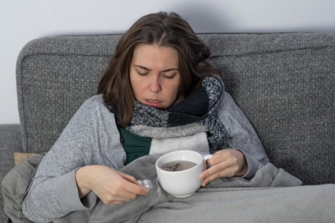 Sick woman covered with a blanket drinking an infusion on the couch in her home