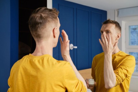 Middle-aged man applying anti-aging cream standing in bedroom in front of the mirror