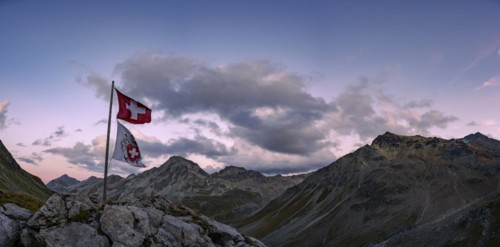 Swiss flag and flag of the SAC with cloudy sky at blue hour and Engadine mountains