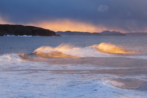 Large waves crash in a winter storm and the swirling spray is illuminated by the warm light of the evening sun