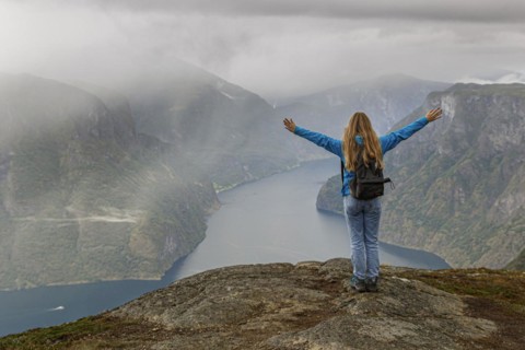 A girl stands on a mountain above the Aurlandsfjord in Norway and stretches her hands up in the air