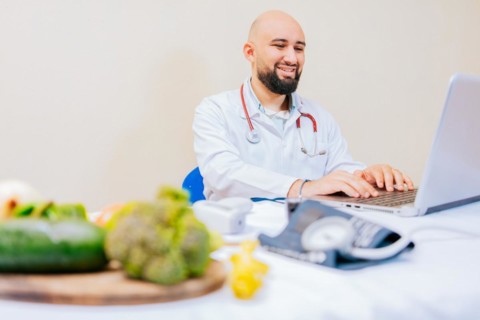 Smiling nutritionist with laptop at desk. Nutritionist doctor using laptop at workplace. Bearded nutritionist doctor working on laptop at desk