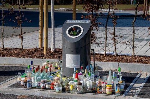 Bottles and jars stand in front of an overcrowded waste glass collection container