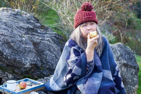 White-haired woman with red cap eating an apple sitting on a rock in the field